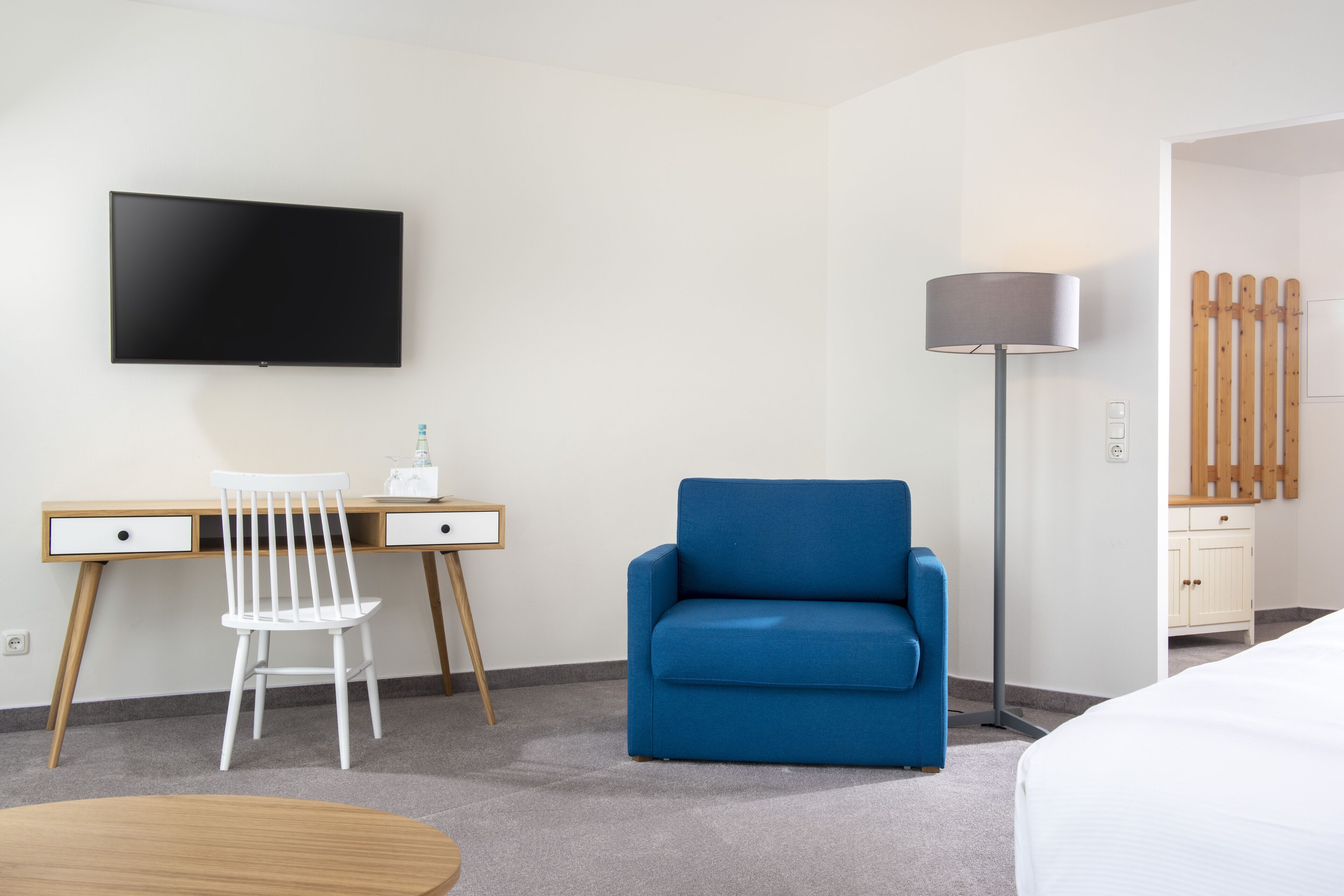 A hotel room with a flat-screen TV mounted on the wall, a desk with a chair, a blue armchair, a floor lamp, and a bed.