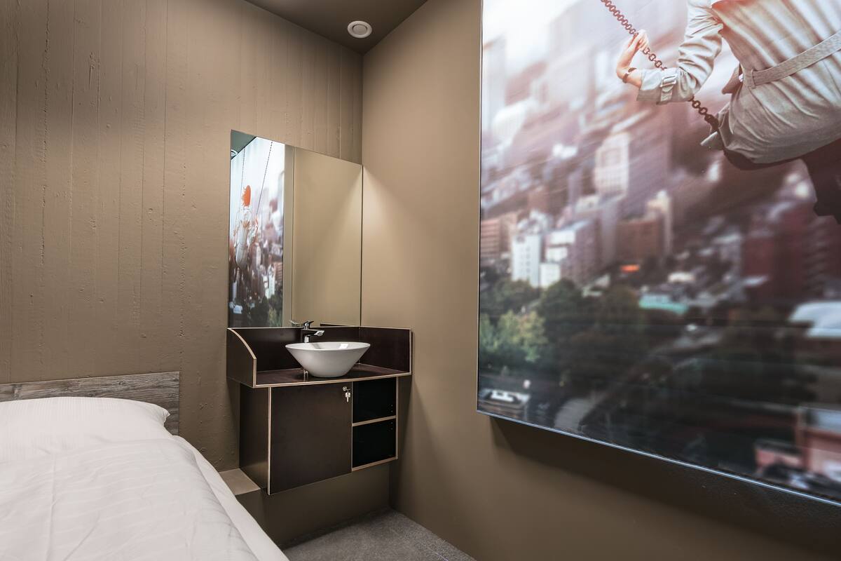 A modern hotel room with a bed, a built-in sink, a mirror, and a large window with a city view.