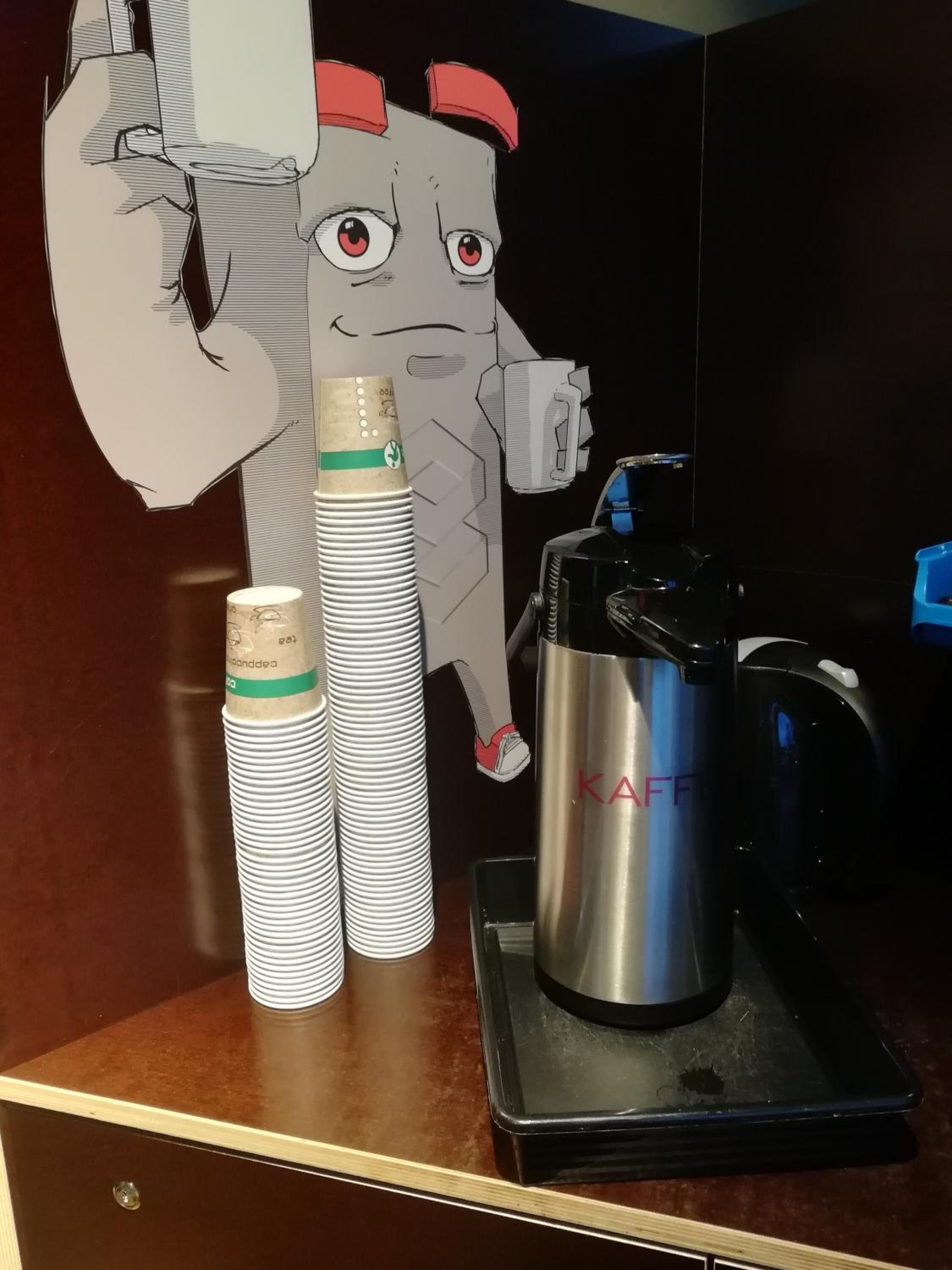 Una encimera de cocina con una cafetera, una pila de vasos desechables y un recorte de un personaje de dibujos animados.