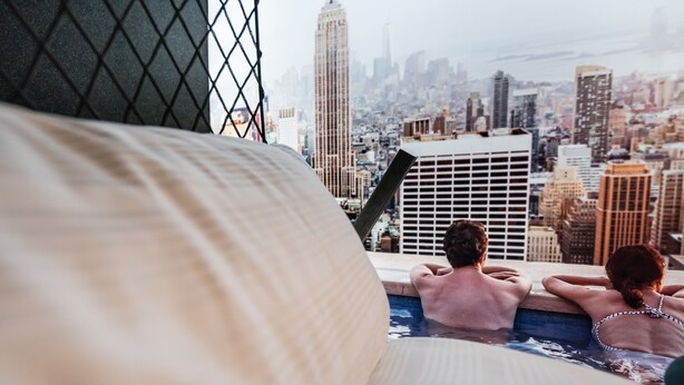 Una pareja relajándose en una piscina en la azotea con vista a un paisaje urbano, incluyendo un rascacielos destacado.