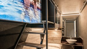 Habitación de hotel moderna con una pantalla grande que muestra un paisaje urbano, una cama con edredón blanco, una mesita de noche con un cuenco blanco y un banco empotrado con escalones que llevan a la cama.