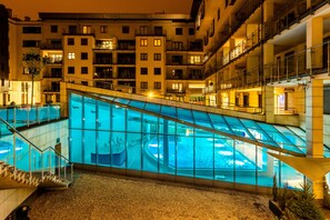 A modern hotel with a glass-enclosed swimming pool, multiple balconies, and a well-lit exterior. - A11 Apartments & SPA Dermique (Kraków)