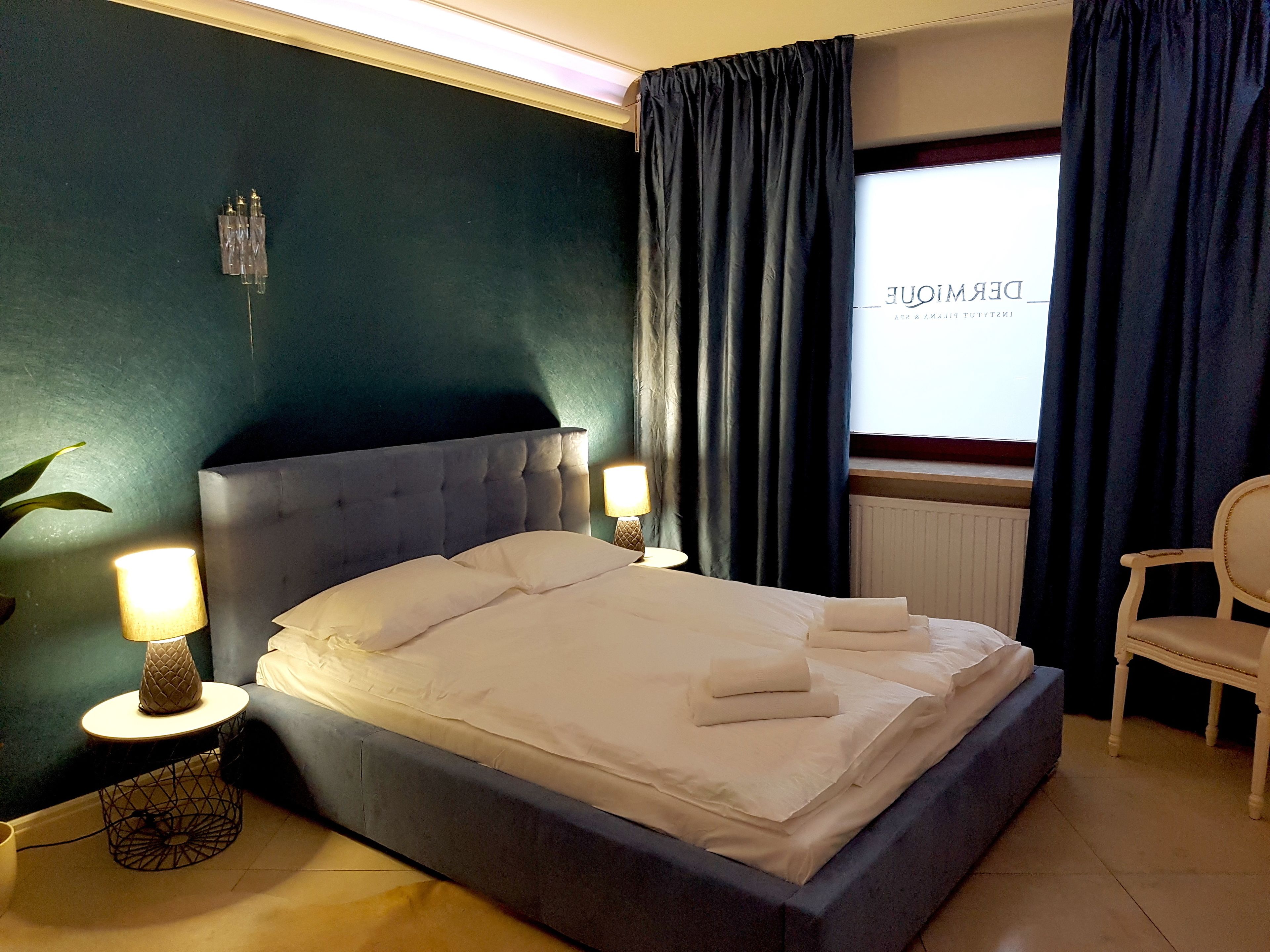 Un letto rifatto con lenzuola bianche, un comodino rotondo con una lampada e una sedia alla destra.