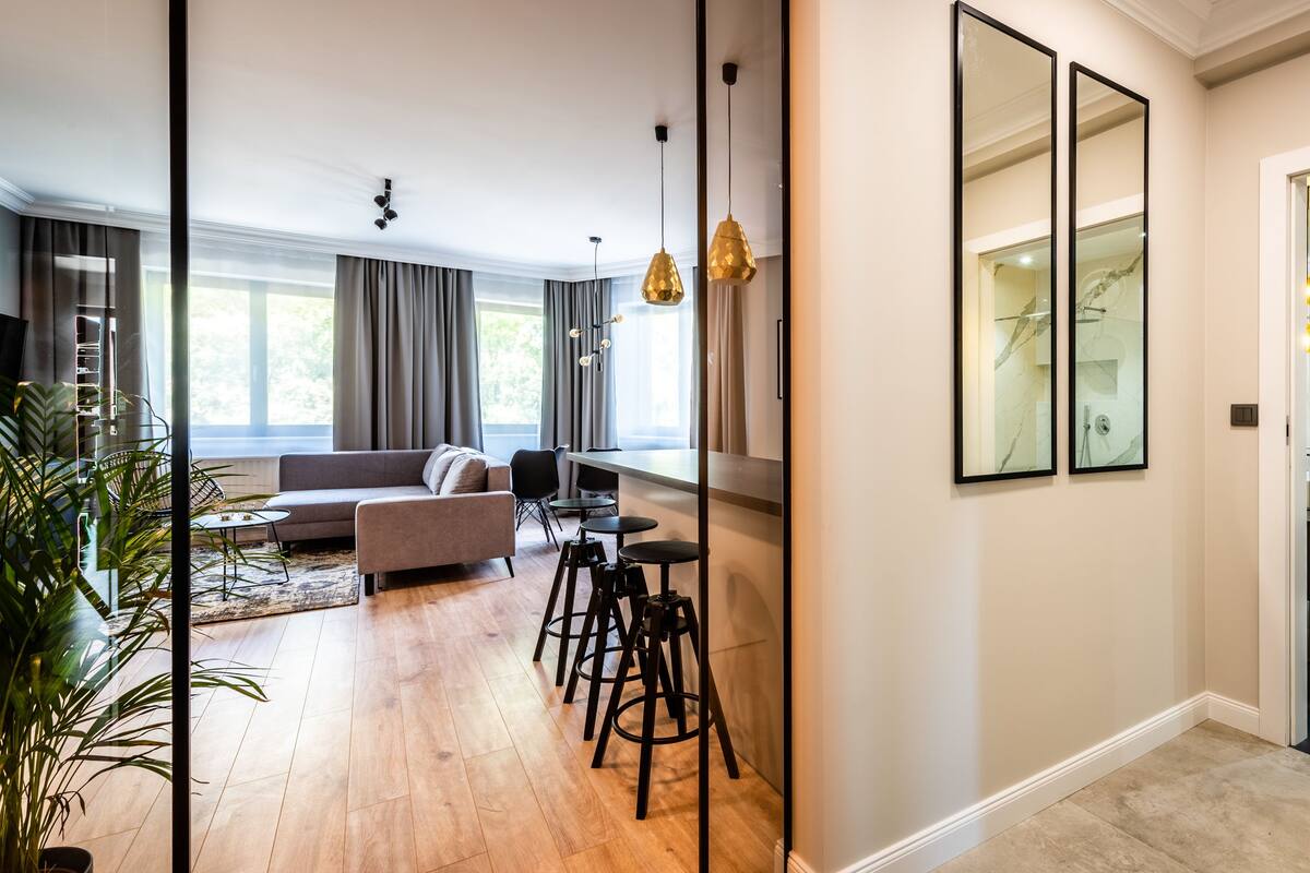 A modern living room with a sofa, coffee table, and bar stools. There is a large window with curtains, a ceiling light, and a mirror on the wall.