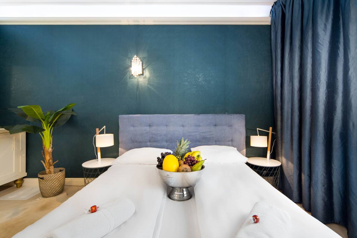 A hotel room with a bed, a fruit bowl on the bed, a potted plant, and two bedside tables with lamps.