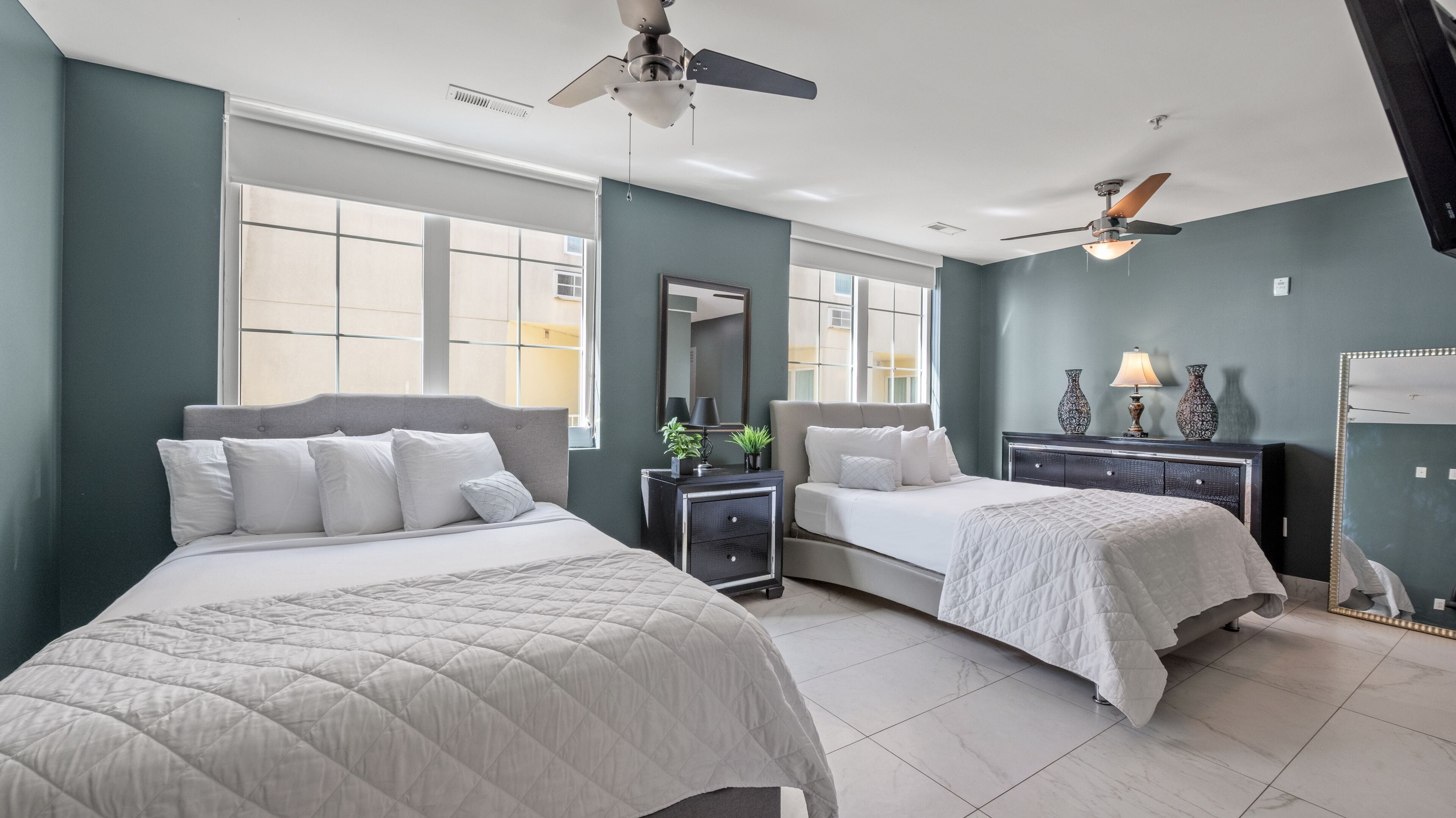 A modern hotel room with two beds, a ceiling fan, large windows, and a nightstand with decorative items.