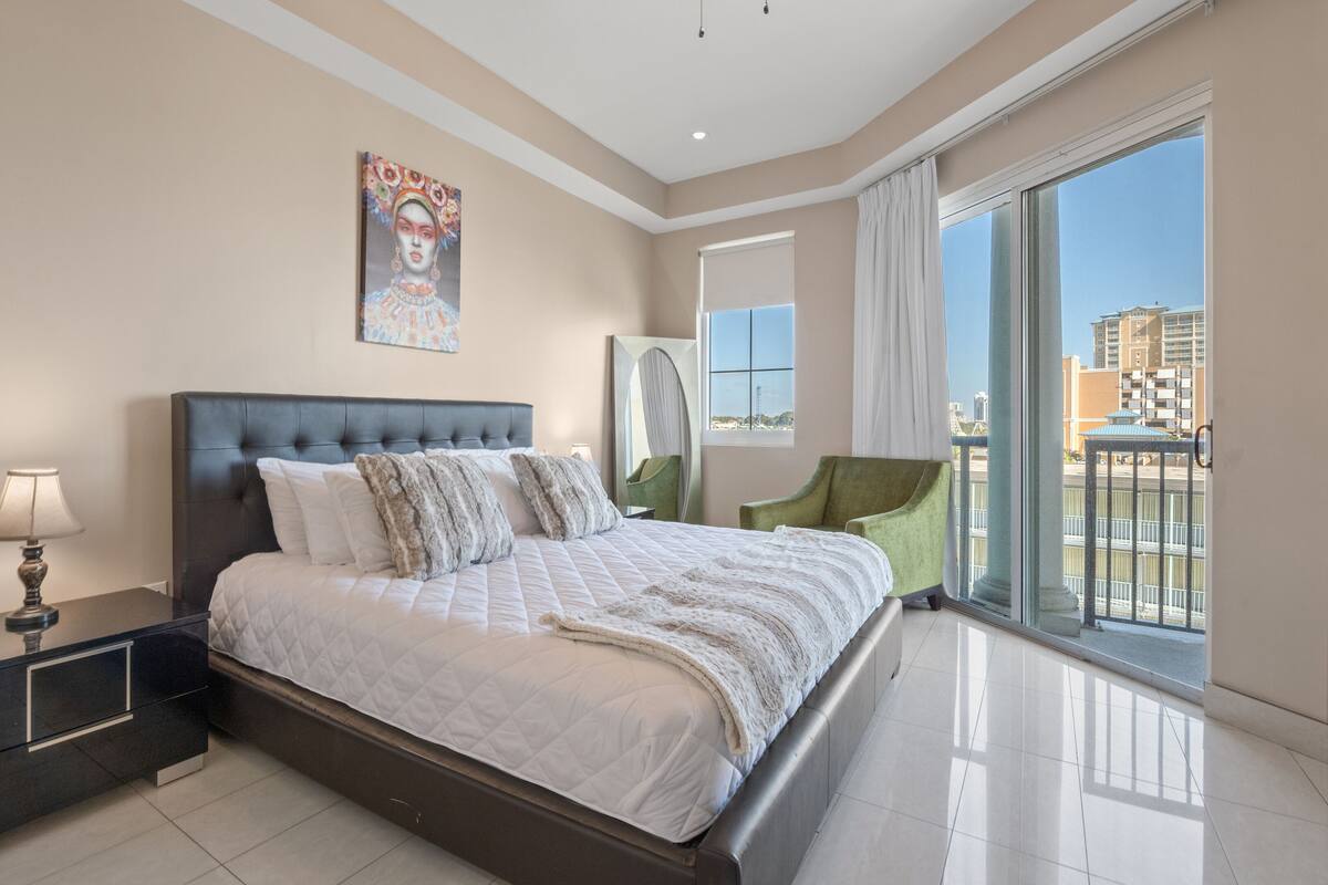 Ein modernes Schlafzimmer mit einem großen Bett, einem Nachttisch, einem grünen Sessel und Blick auf die Stadt vom Balkon aus.