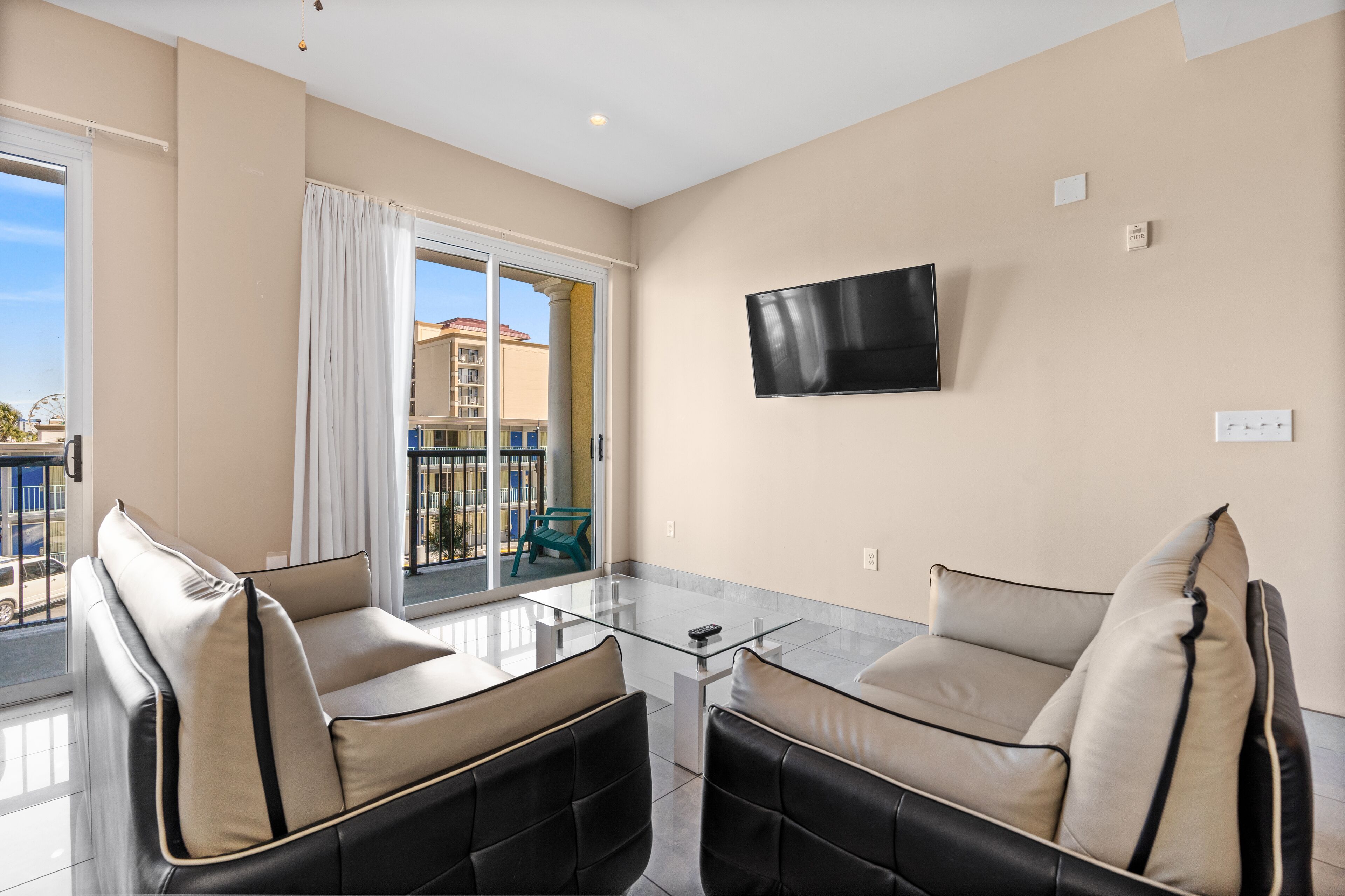 A modern living room with a glass coffee table, two leather sofas, a flat-screen TV, and a balcony with a view of buildings.