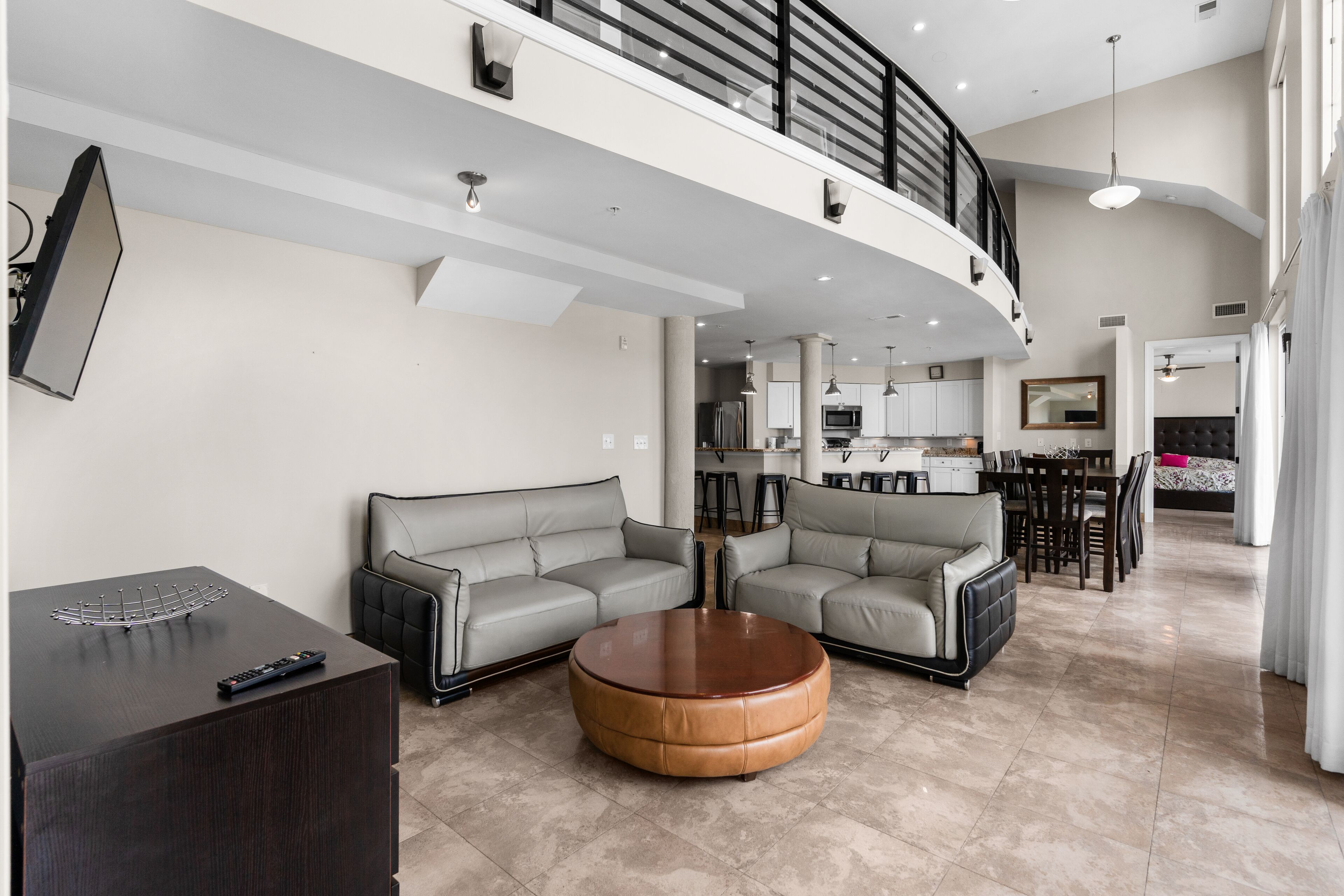 A spacious living area with a high ceiling, a round coffee table, two grey sofas, a dark wooden side table, and a kitchen in the background.