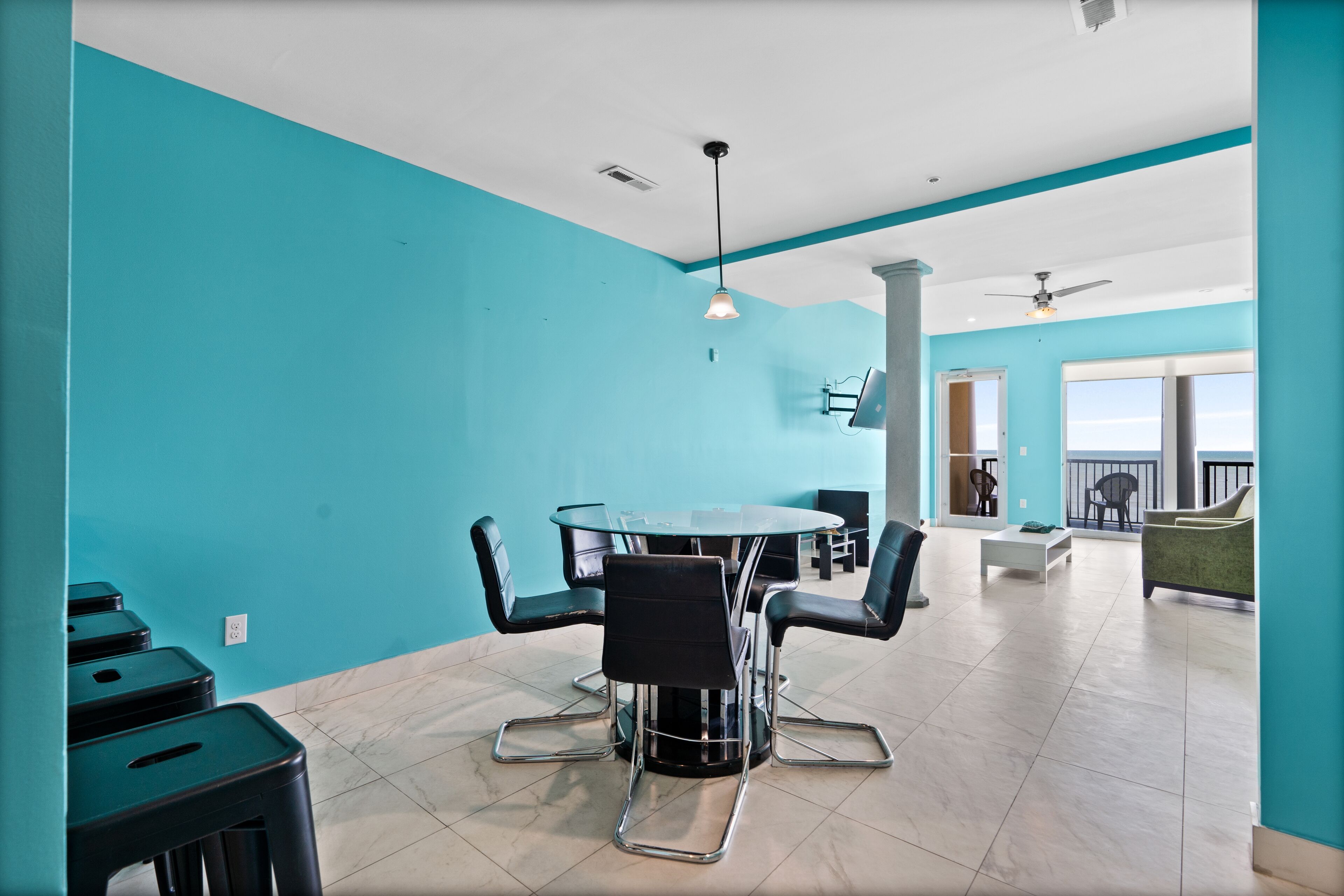 A modern room with teal walls, a round table with chairs, a ceiling fan, and a balcony with a view.
