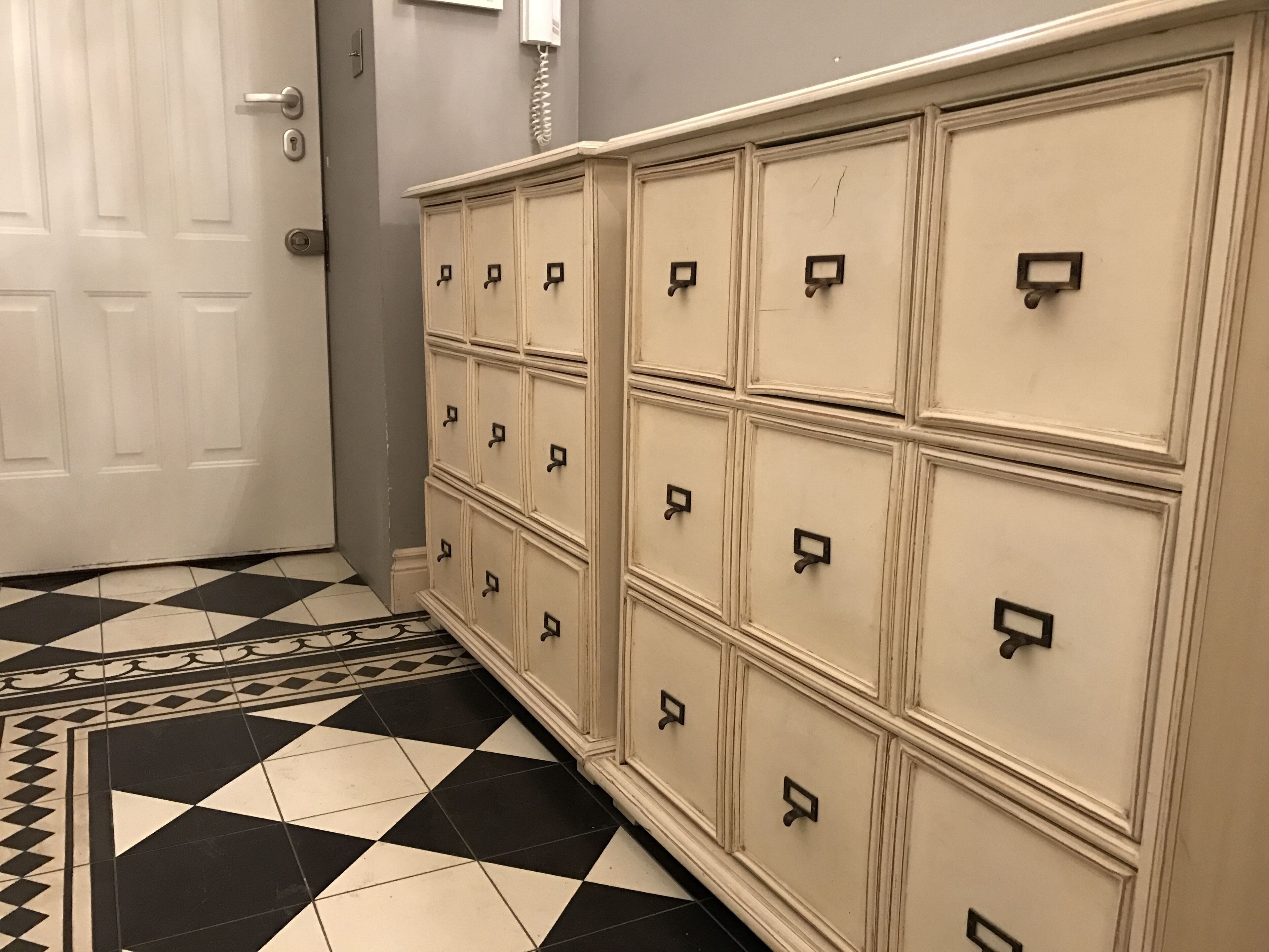 A hallway with a checkered floor, a white door, and a row of cream-colored storage cabinets with black handles.