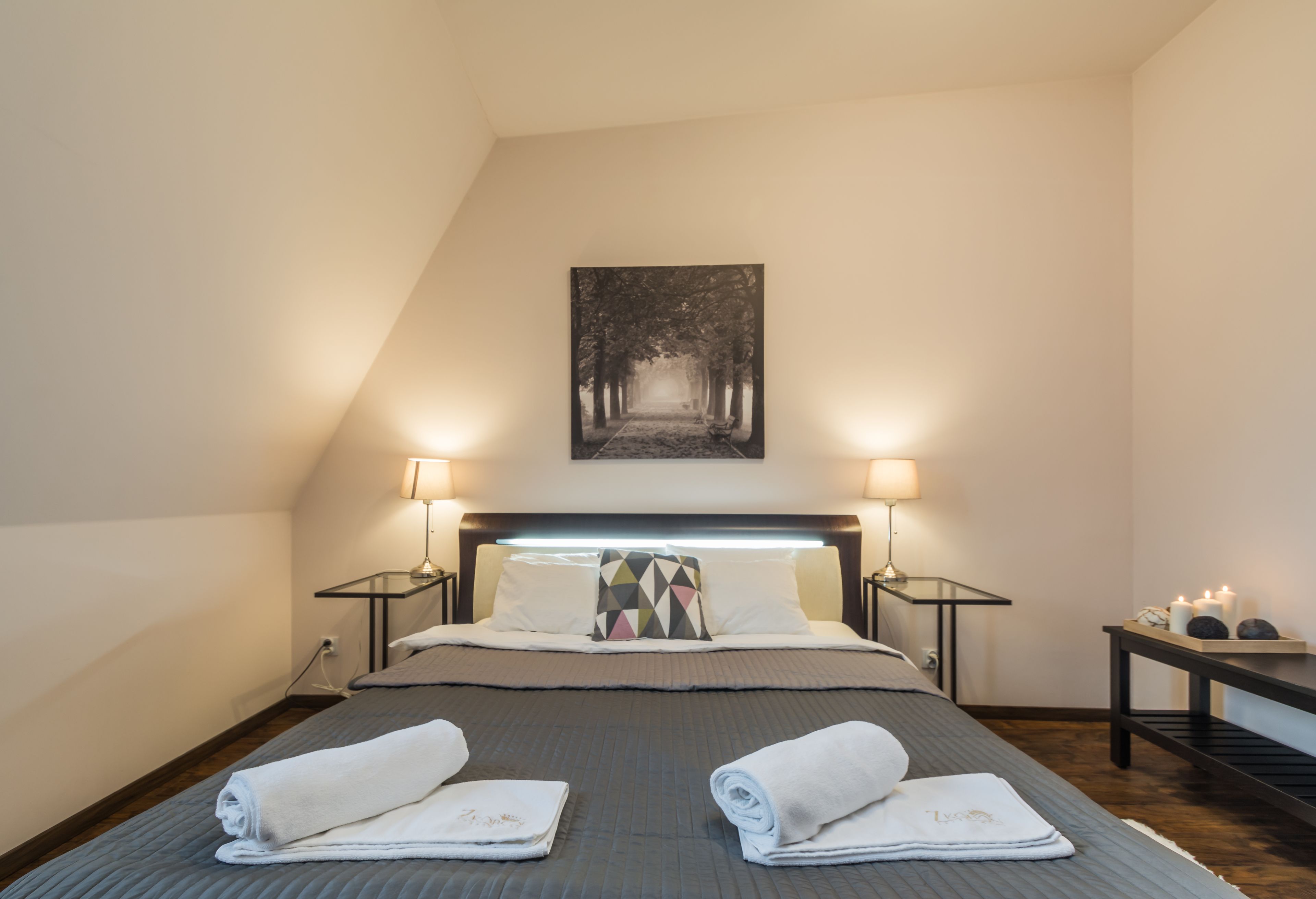 A neatly made bed with two rolled towels, a bedside table with a lamp, and a wall-mounted picture.