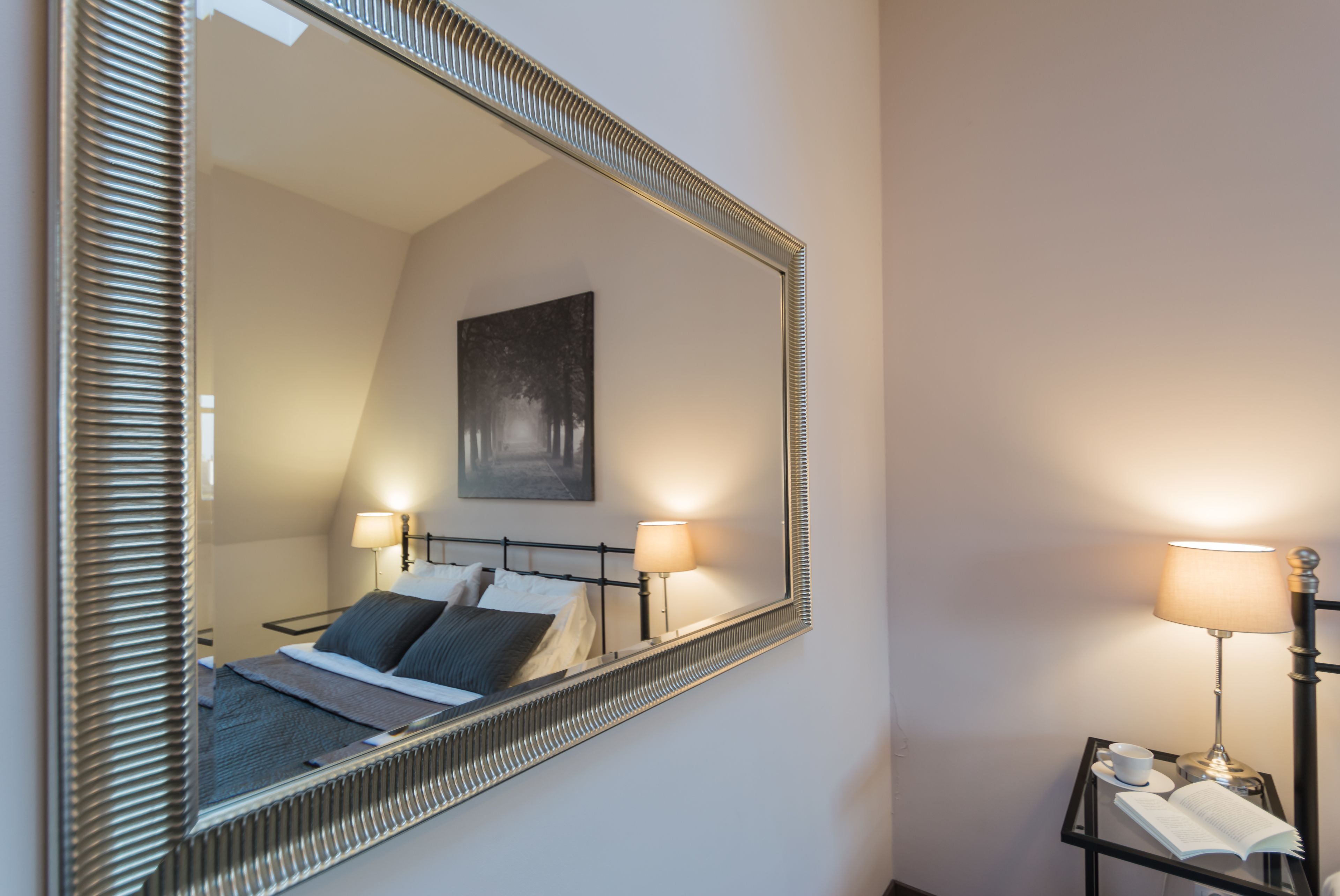 A neatly made bed with pillows, a bedside lamp, a small table with a cup and book, and a framed mirror on the wall.