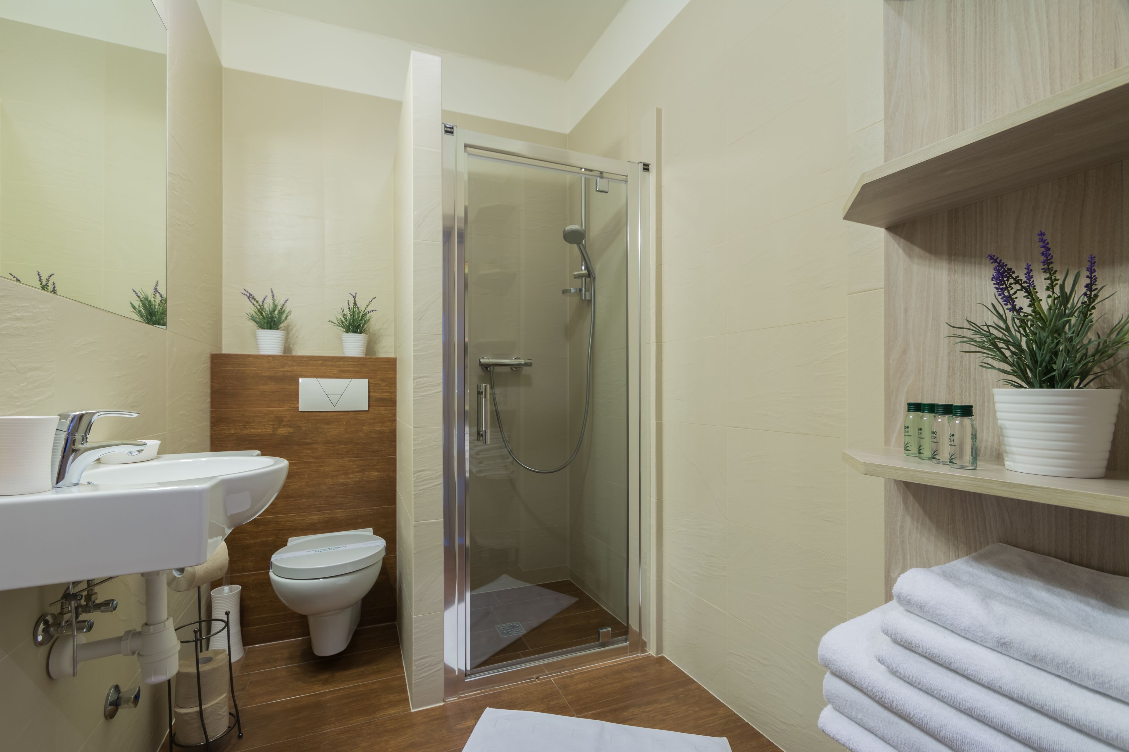 A modern bathroom with a glass-enclosed shower, a white sink, and a toilet. There are potted plants on the shelves and a towel rack with towels.