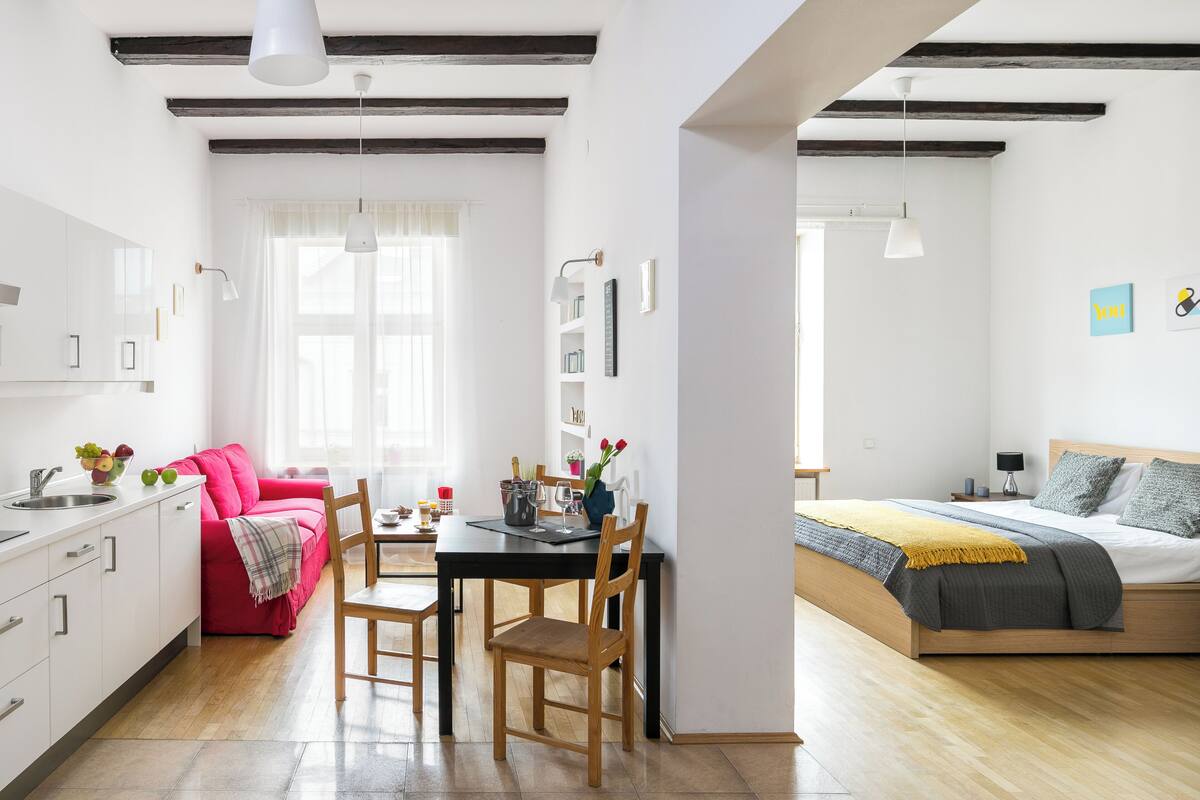 Un apartamento moderno con cocina, comedor y dormitorio. La cocina tiene armarios blancos, una mesa de comedor con sillas y un sofá rojo. El comedor tiene una mesa con un jarrón de flores. El dormitorio tiene una cama con una manta y almohadas grises.