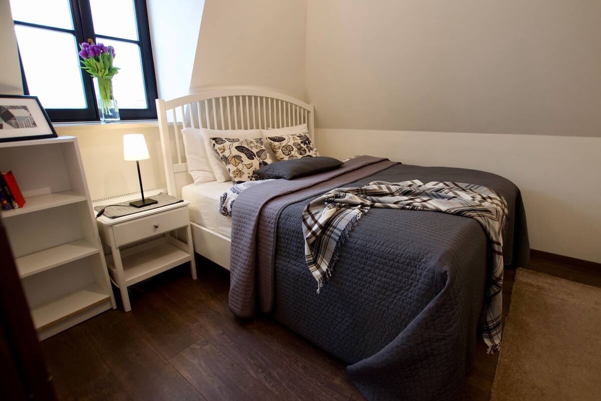 Un dormitorio ordenado con cama, mesita de noche, lámpara, y una pequeña estantería con libros y un jarrón de flores.