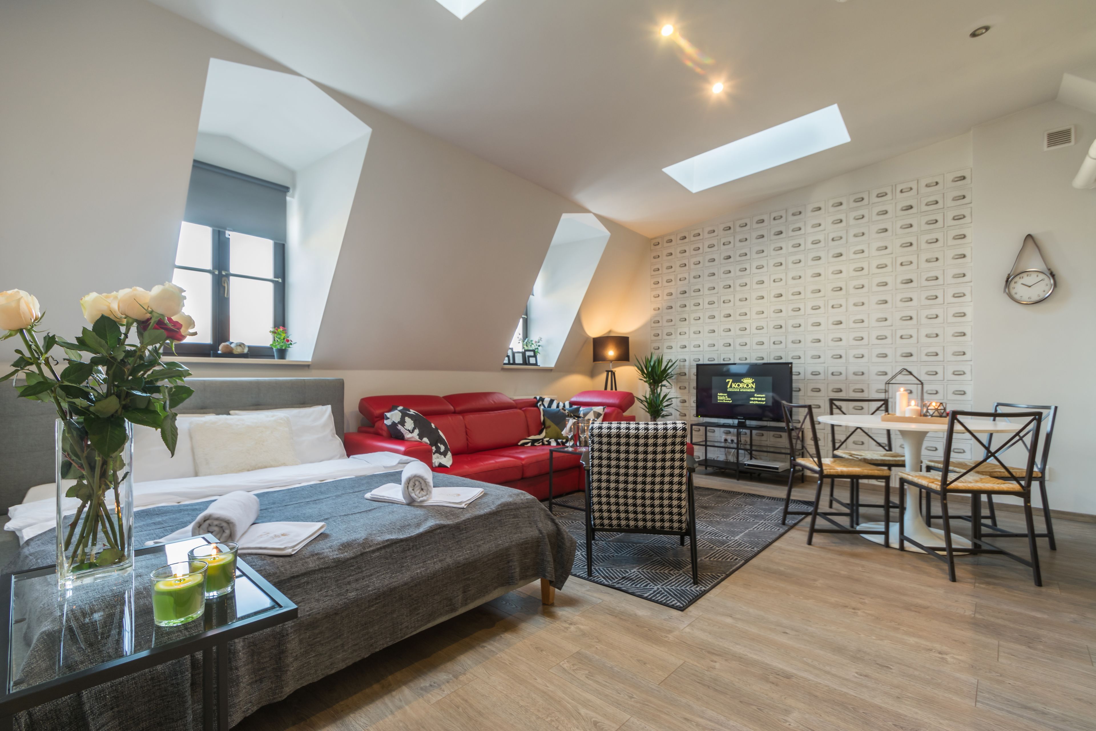 Quarto de hotel moderno com cama, sofá vermelho, mesa de jantar com cadeiras, televisão e uma grande janela com claraboias.