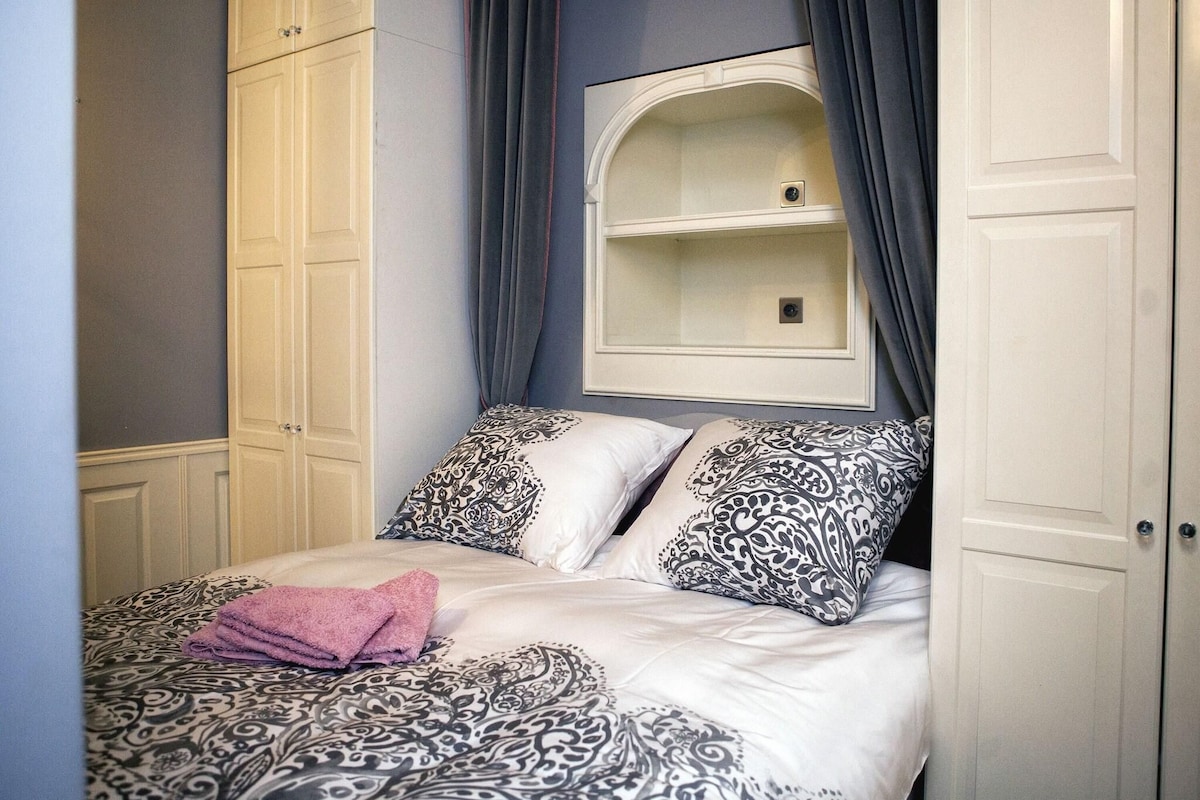 A bedroom with a bed, patterned bedding, a shelf above the bed, and a wardrobe.