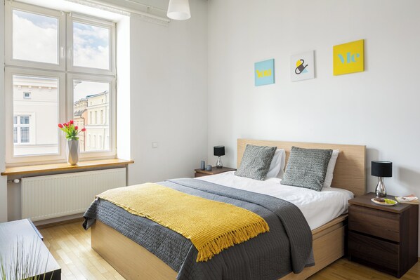 A modern bedroom with a large bed, a bedside table, a nightstand, a window with a view of buildings, and artwork on the wall. - 7 Koron - Exclusive Apartments (Kraków)
