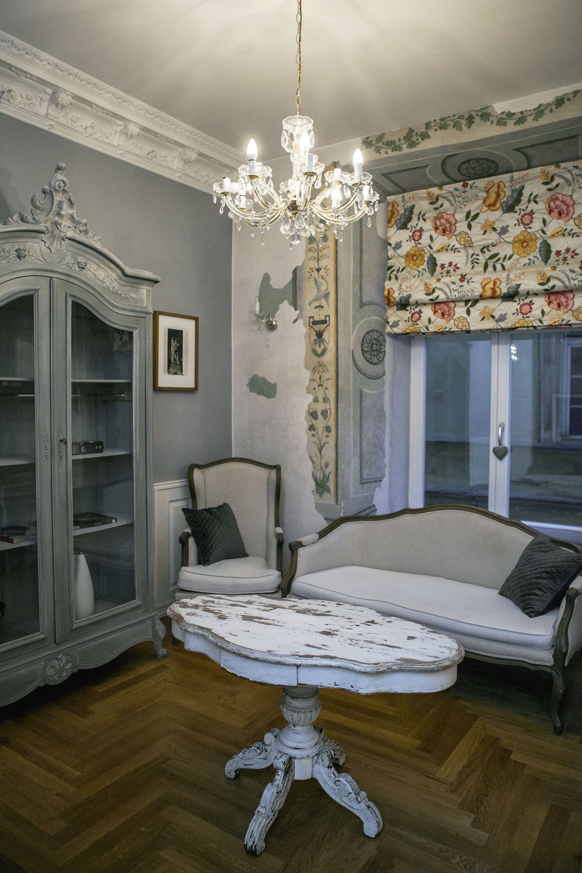 Une pièce agrémentée d’un lustre, d’une lambrequin à motifs floraux, d’un canapé blanc, d’un fauteuil blanc et d’une commode ancienne.