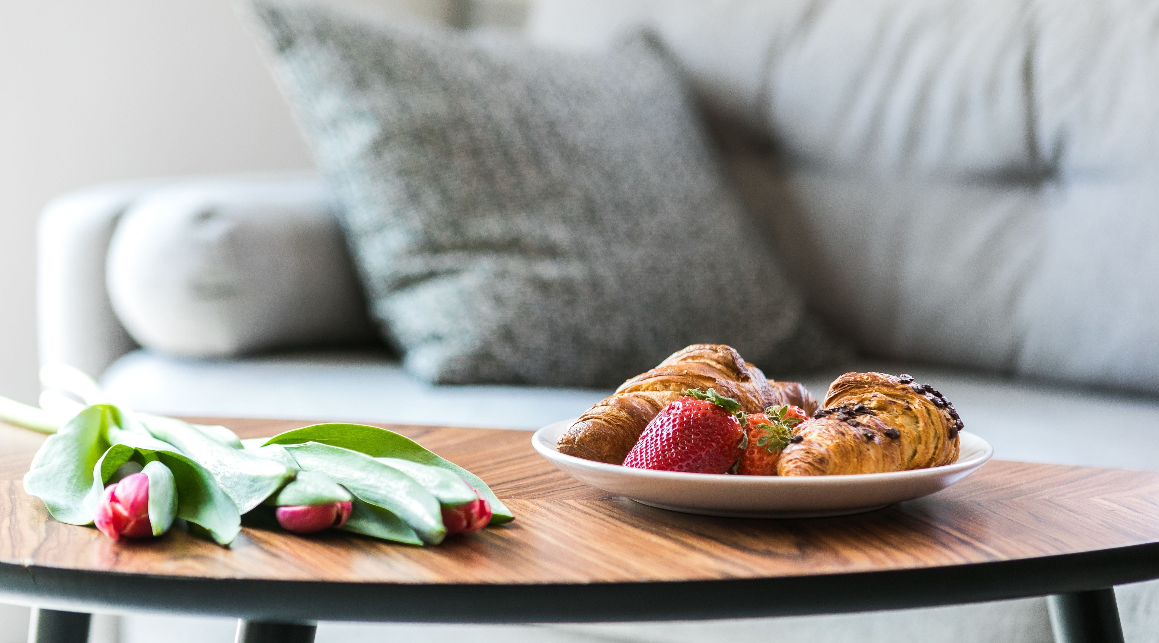 A plate of croissants and strawberries on a round table with a wooden surface, in front of a grey sofa with a pillow.