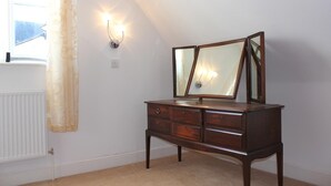 A wooden dresser with a mirror and two wall sconces in a room with a sloped ceiling and a window with curtains. - George Hotel (Bishop's Stortford)