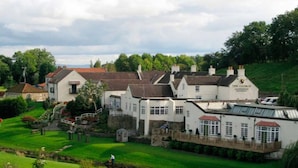 A traditional white building with a red roof, likely a pub or inn, situated beside a river with a well-maintained grassy area and a small bridge. - George Hotel (Bishop's Stortford)