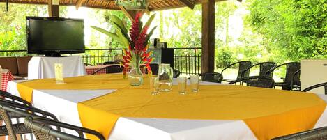 Un área de comedor al aire libre con techo de paja, una mesa grande cubierta con mantel amarillo y sillas de mimbre.