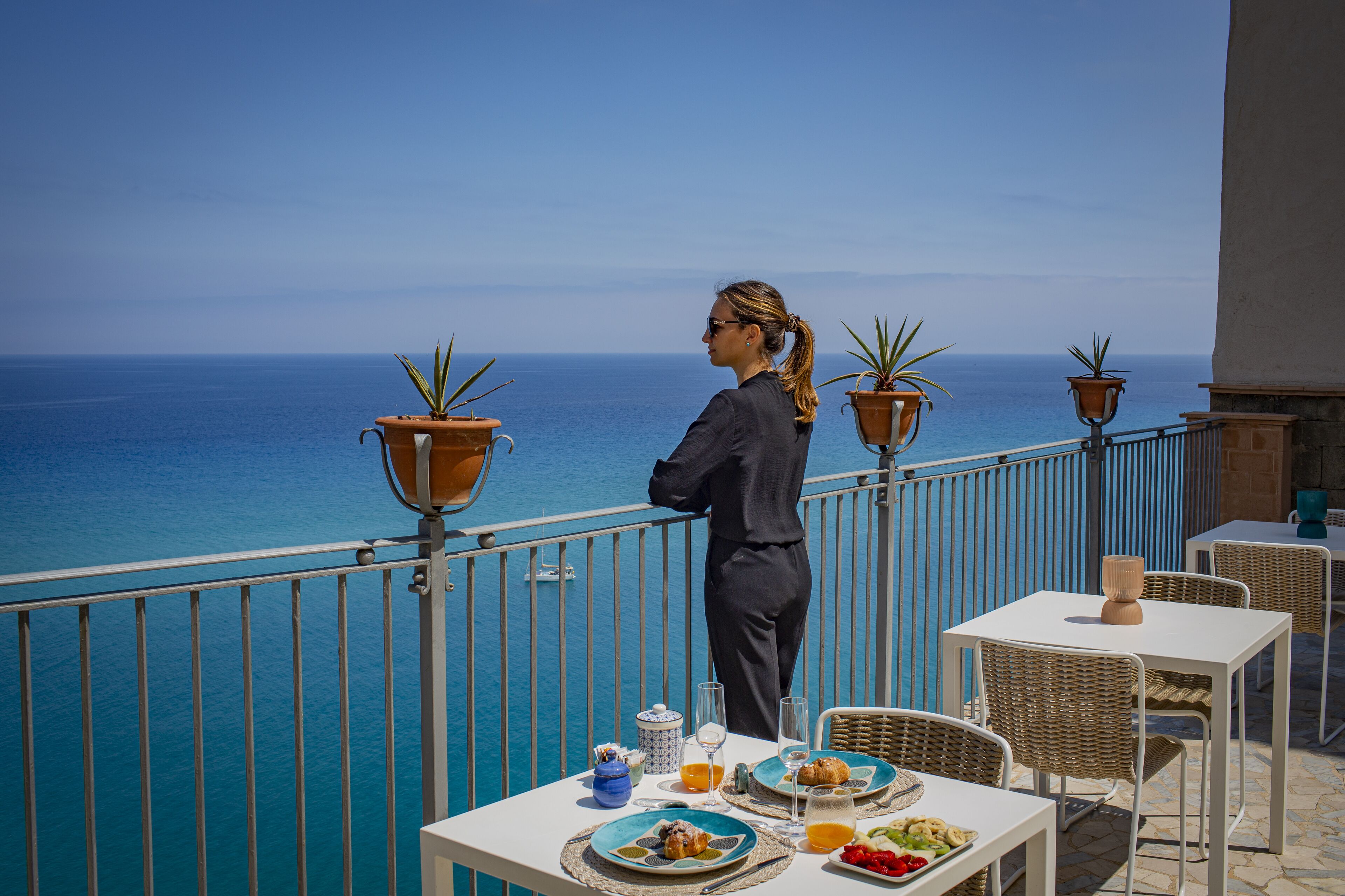A balcony with a view of the sea, a table with breakfast items, and potted plants.
