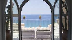 Vista a través de un amplio ventanal hacia un balcón con sofá blanco y plantas en macetas, con vistas al mar.
