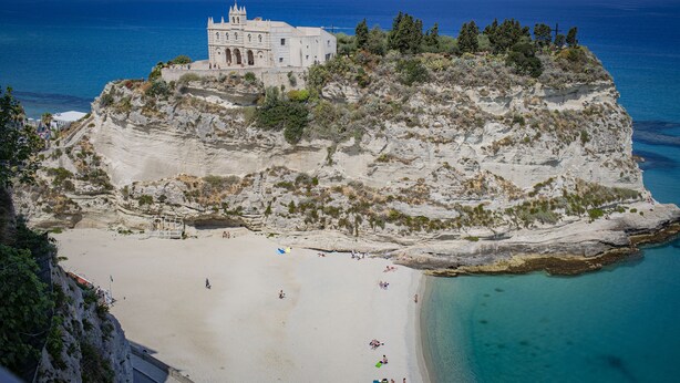 Un edificio costero de fachada blanca, asentado sobre un acantilado con vistas a una playa.