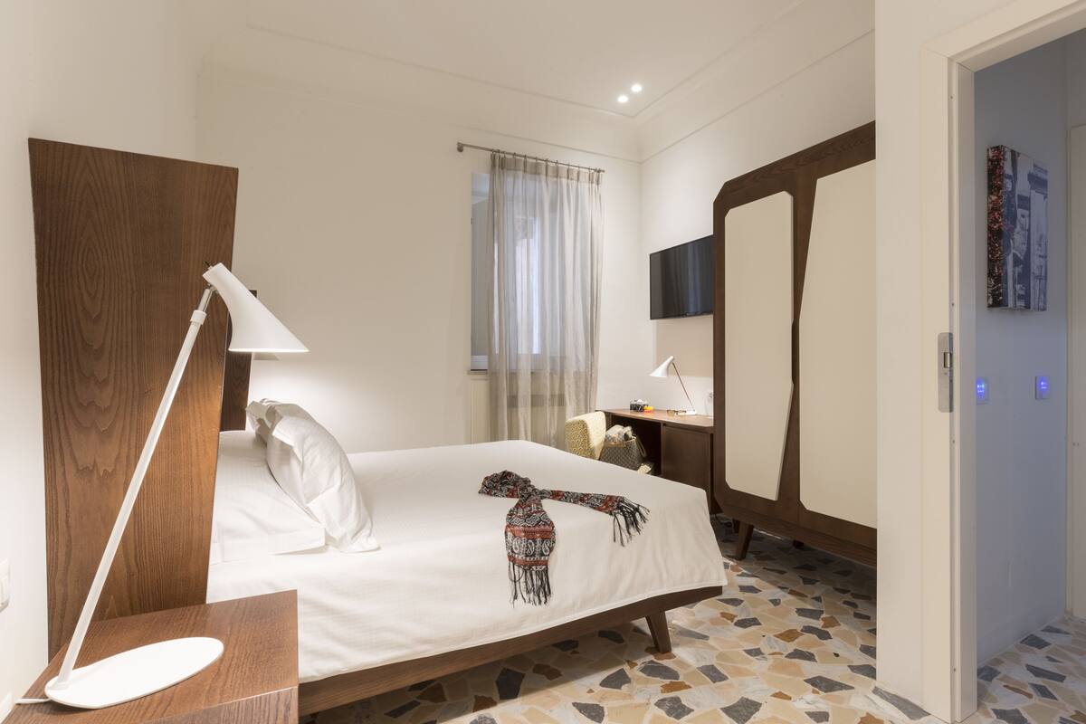 A hotel room with a bed, a desk, a chair, a mirror, and a window with curtains.