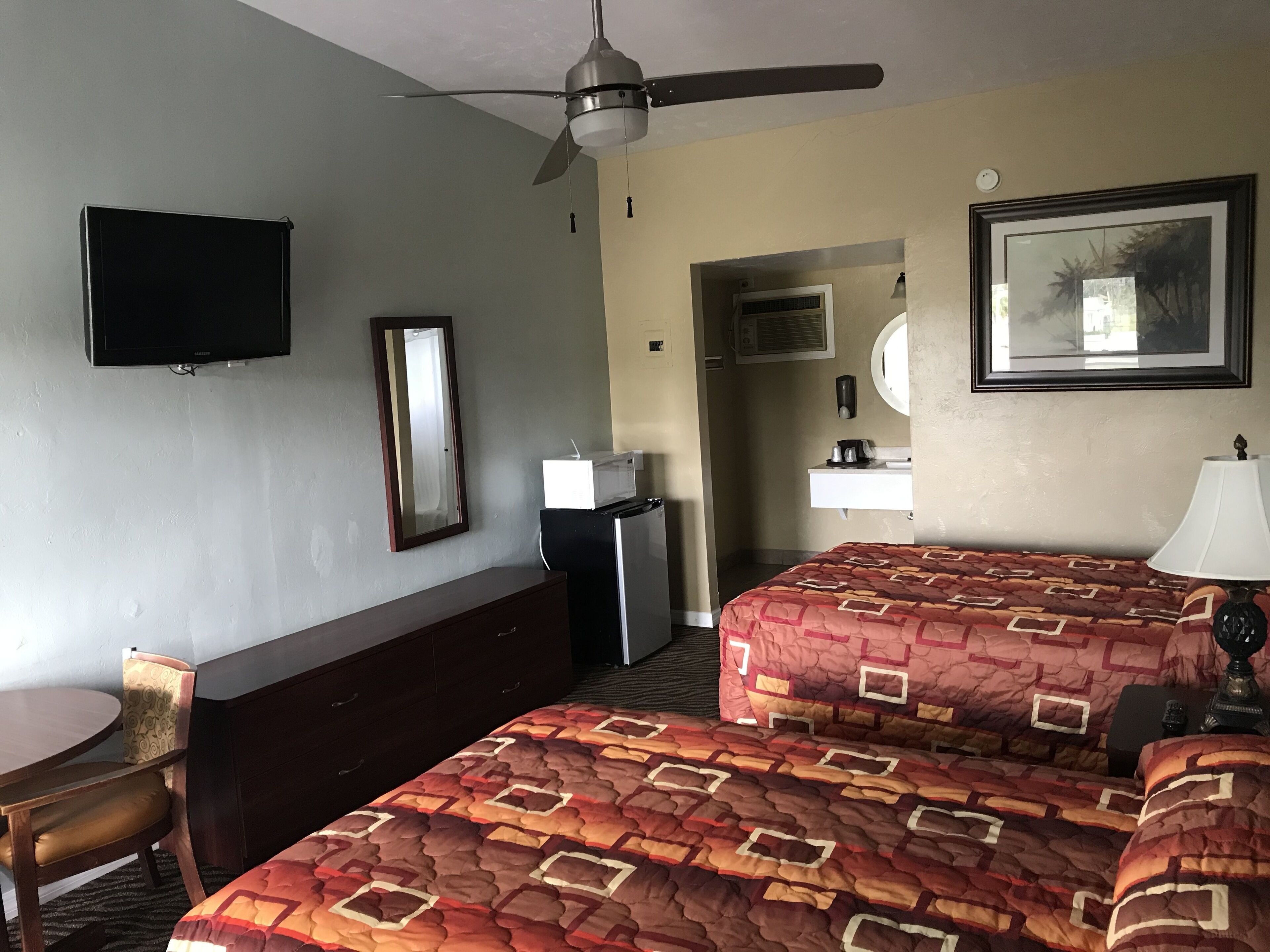A hotel room with two beds, a desk, a chair, a television, a ceiling fan, a mirror, a framed picture, and a small kitchenette.