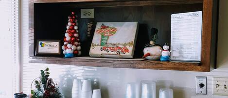 A kitchen area with a wooden shelf displaying Christmas decorations, a framed sign for 'Mistletoe Motel', and a snowman figurine. Below, there are stacks of plastic cups and a tray with snacks.