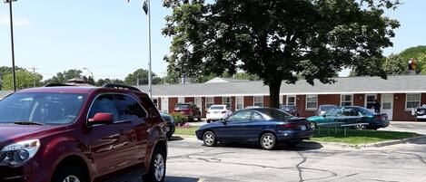 A parking lot with several cars, a large tree, and a building with a red roof in the background.