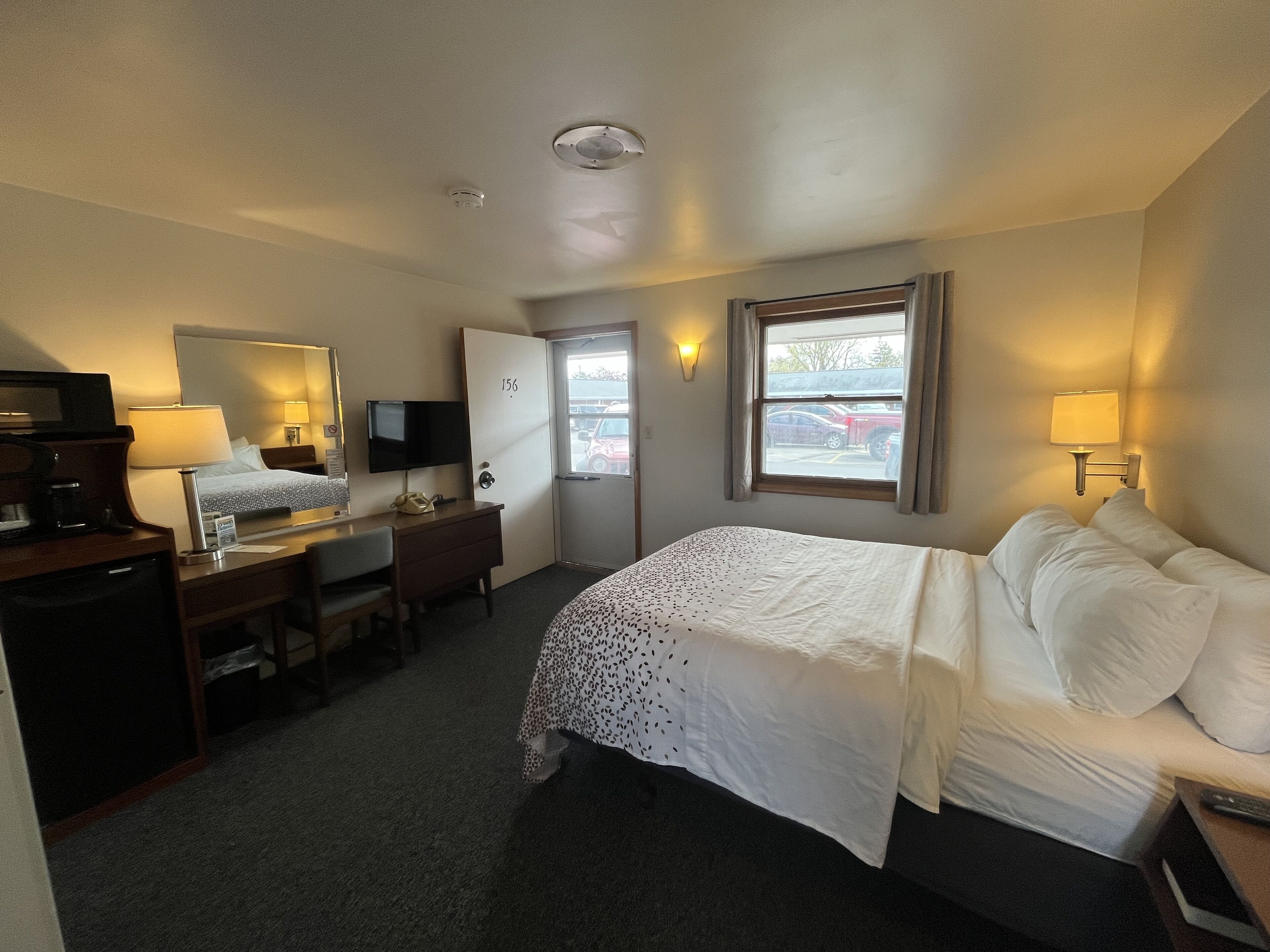 A hotel room with a bed, desk, chair, television, and a window with a view of parked cars outside.