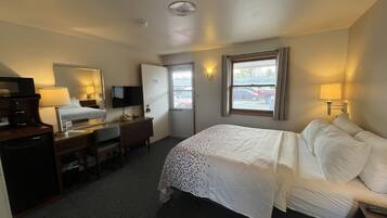 A hotel room with a bed, desk, chair, television, and a window with a view of parked cars outside.