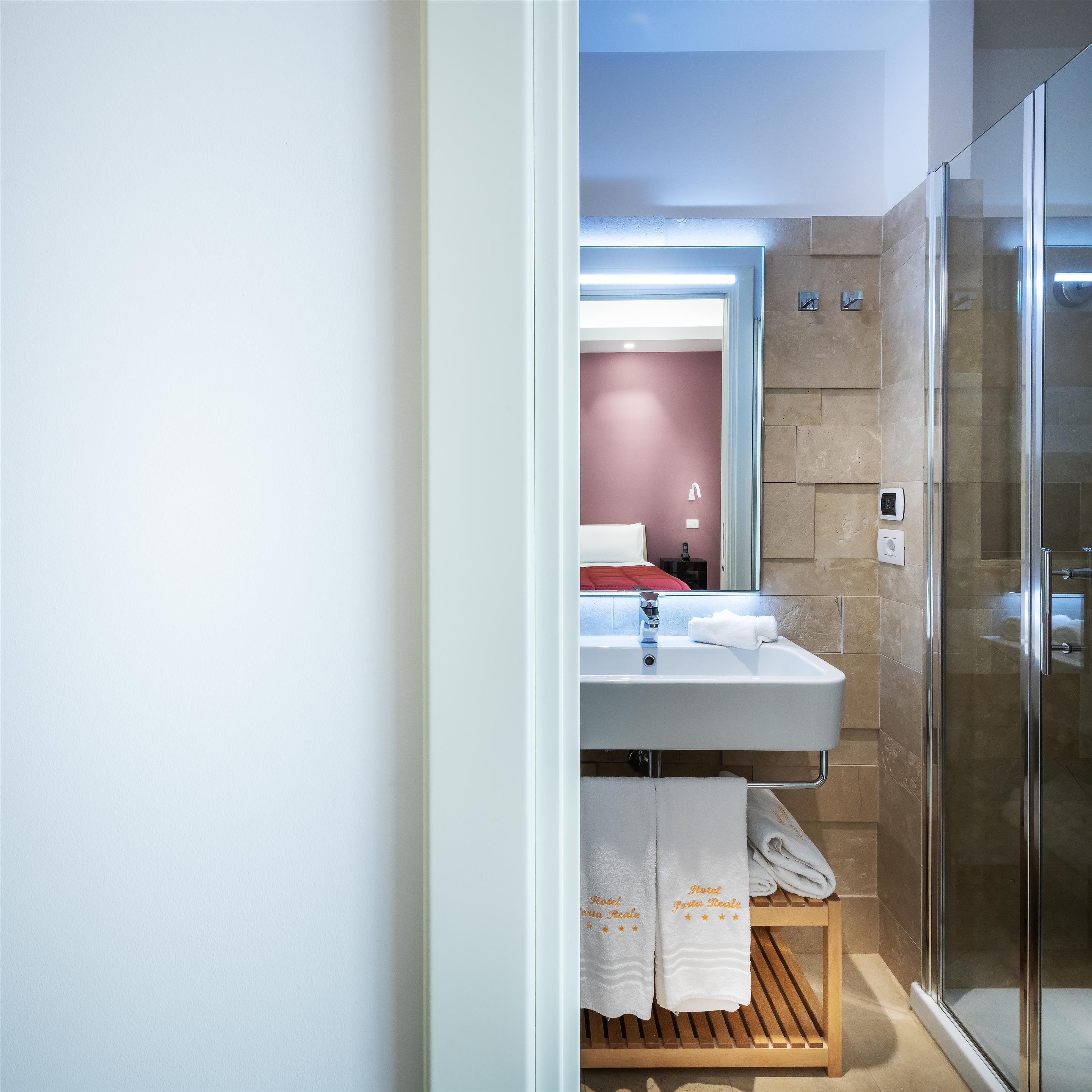 Ein modernes Badezimmer mit Duschkabine aus Glas, einem weißen Waschbecken und einem Spiegel. Auf einem Holzregal liegen Handtücher und im Hintergrund ist eine rote Bettdecke zu sehen.
