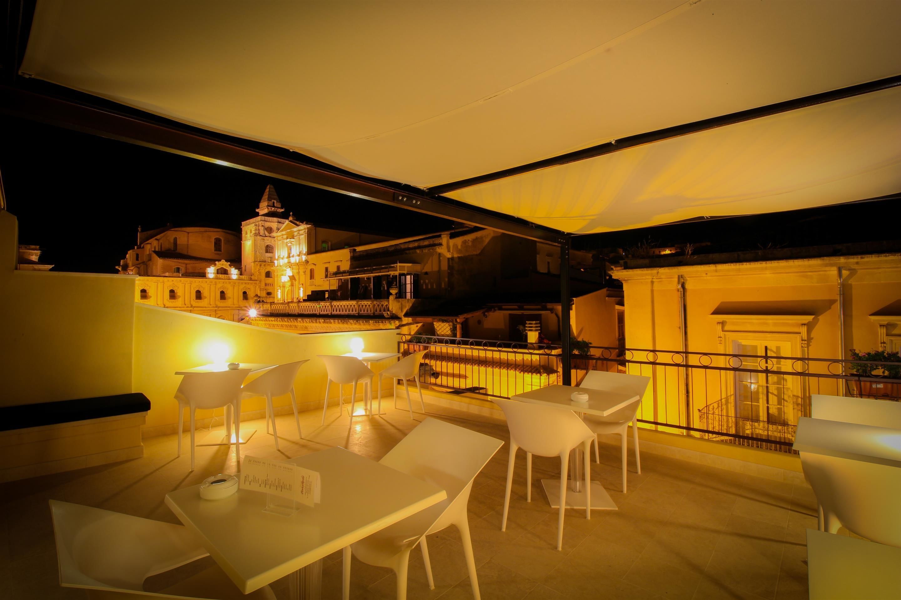 An outdoor dining area with white tables and chairs, a covered section, and a view of a historic building with a bell tower in the background.