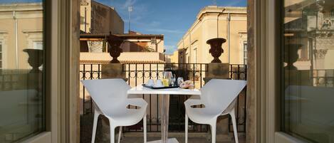Un balcón con dos sillas blancas y una mesa, una pequeña bandeja con objetos y vistas a edificios al fondo.