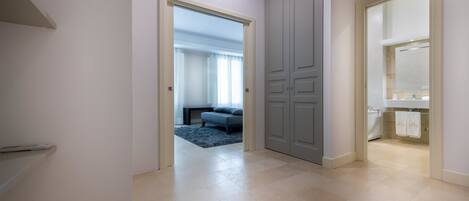 Un pasillo con una puerta que da a un dormitorio con cama y un tapete azul, y otra puerta que conduce a un baño con lavamanos y toallas.
