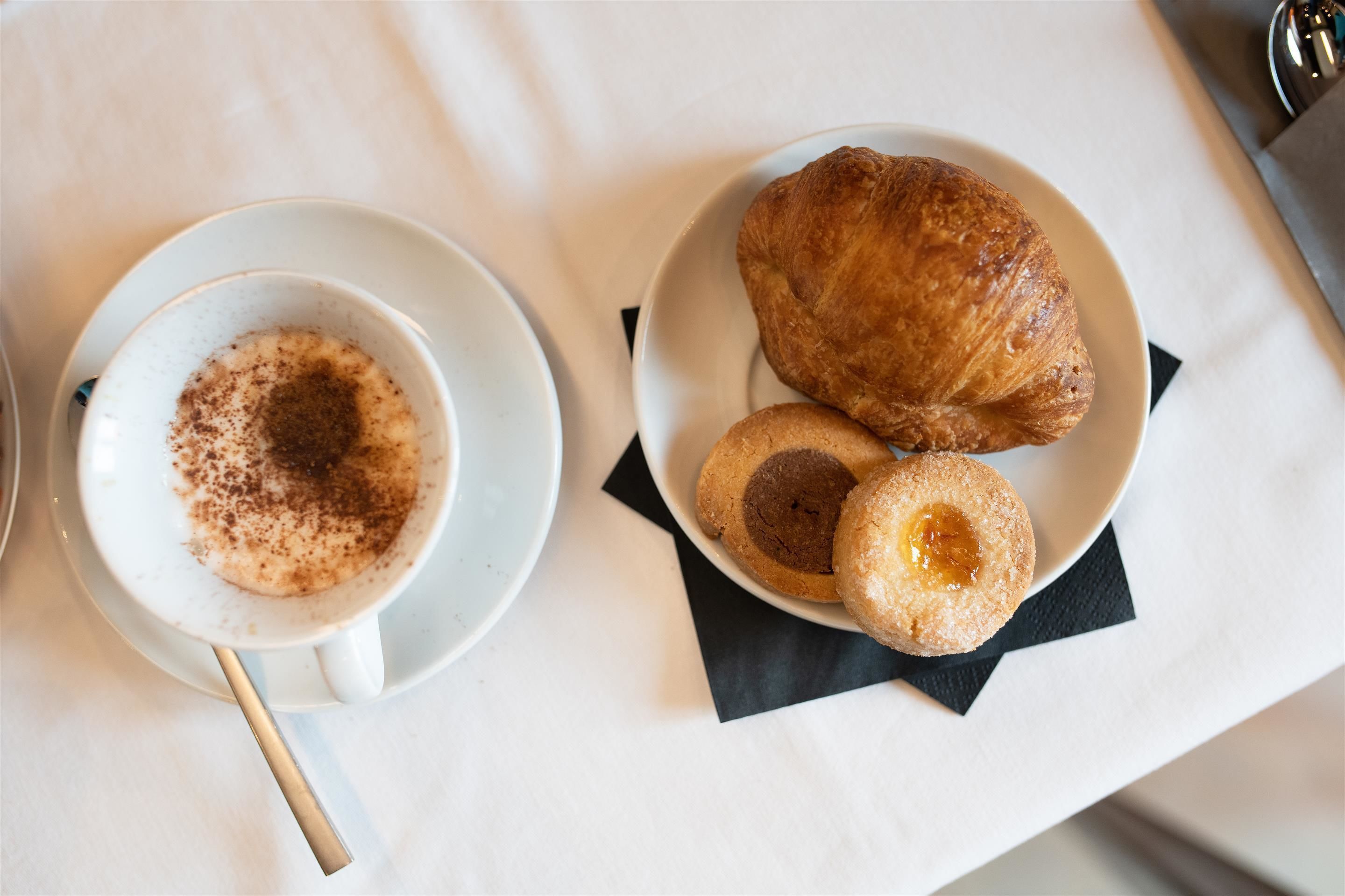 Un tavolo con tovaglia bianca, una tazza di caffè con un cucchiaino, e un piatto con un croissant e due biscotti.