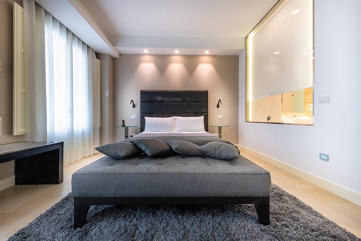 A modern bedroom with a large bed, bedside tables, a bench, and a mirror on the wall.
