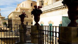 A table with a green glass object, a railing, and a view of historic buildings with balconies and flower pots.