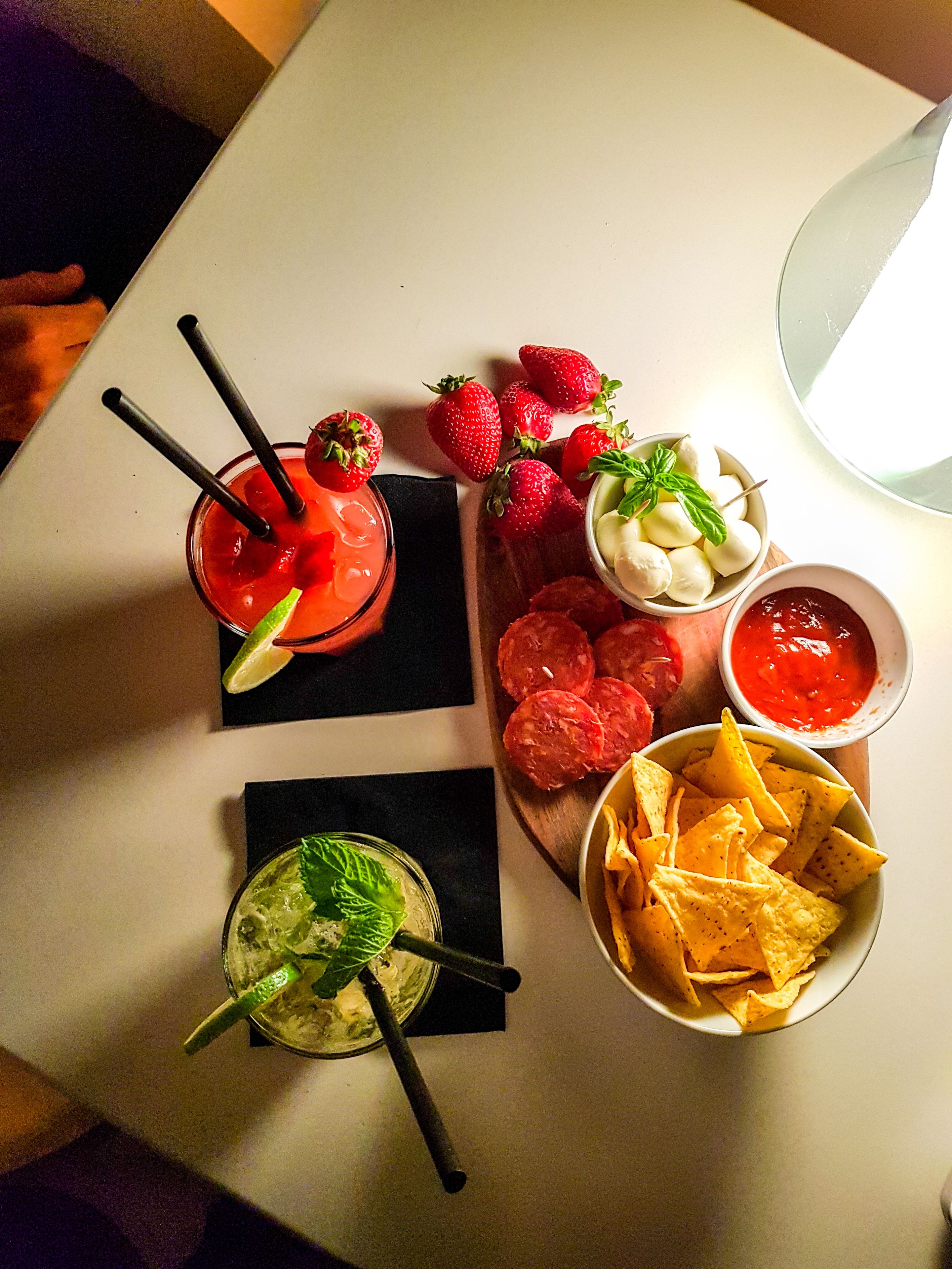 A table with two cocktails, a bowl of nachos, sliced meat, and a plate of strawberries.