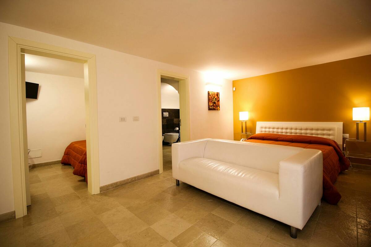 A hotel room with a white sofa, two beds with orange bedding, a TV on the wall, and two bedside lamps.