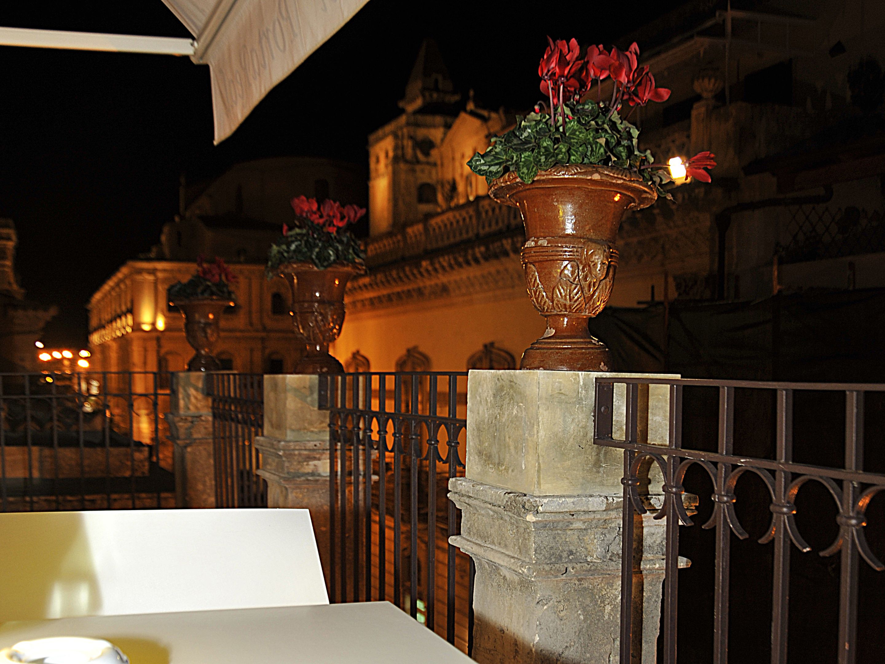 A nighttime view of a historic building with ornate architecture, featuring two large flower pots on pedestals and a metal railing in the foreground.