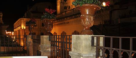 Vista nocturna de un edificio histórico con arquitectura ornamentada, que presenta dos macetas grandes sobre pedestales y una barandilla metálica en primer plano.