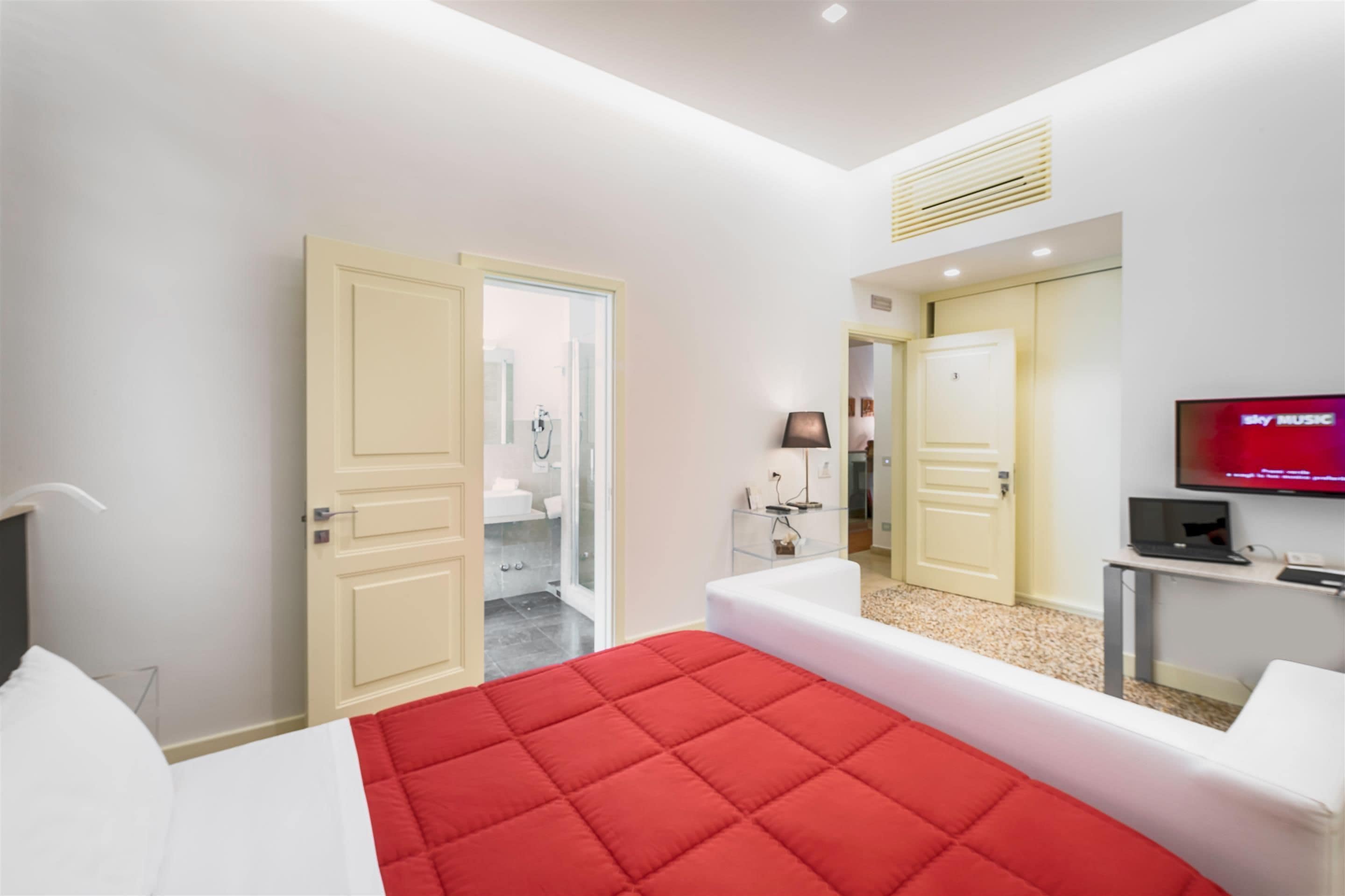 A modern hotel room with a large bed, a TV, a desk, and a bathroom visible through an open door.