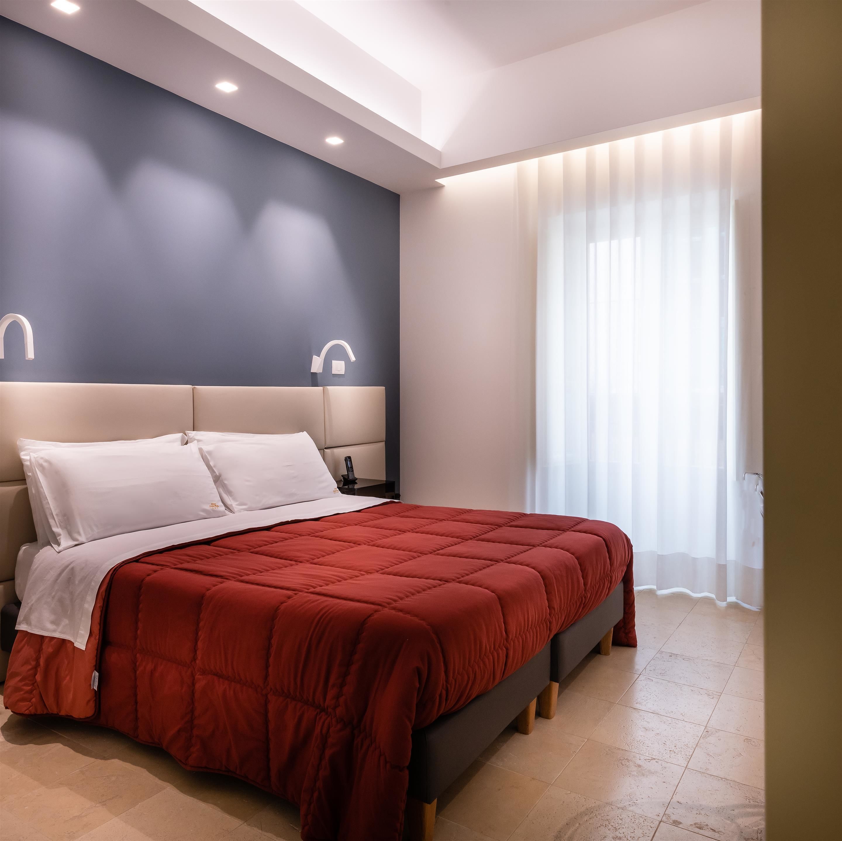 A modern bedroom with a large bed, a headboard with integrated lighting, a bedside table with a phone, and a window with sheer curtains.